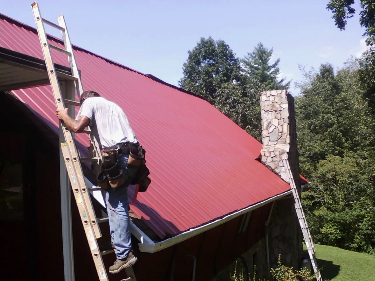 Roofer on a ladder beside a red metal roof and stone chimney — Soffit & Fascia Repair by Roofing Brothers in Lake Helen